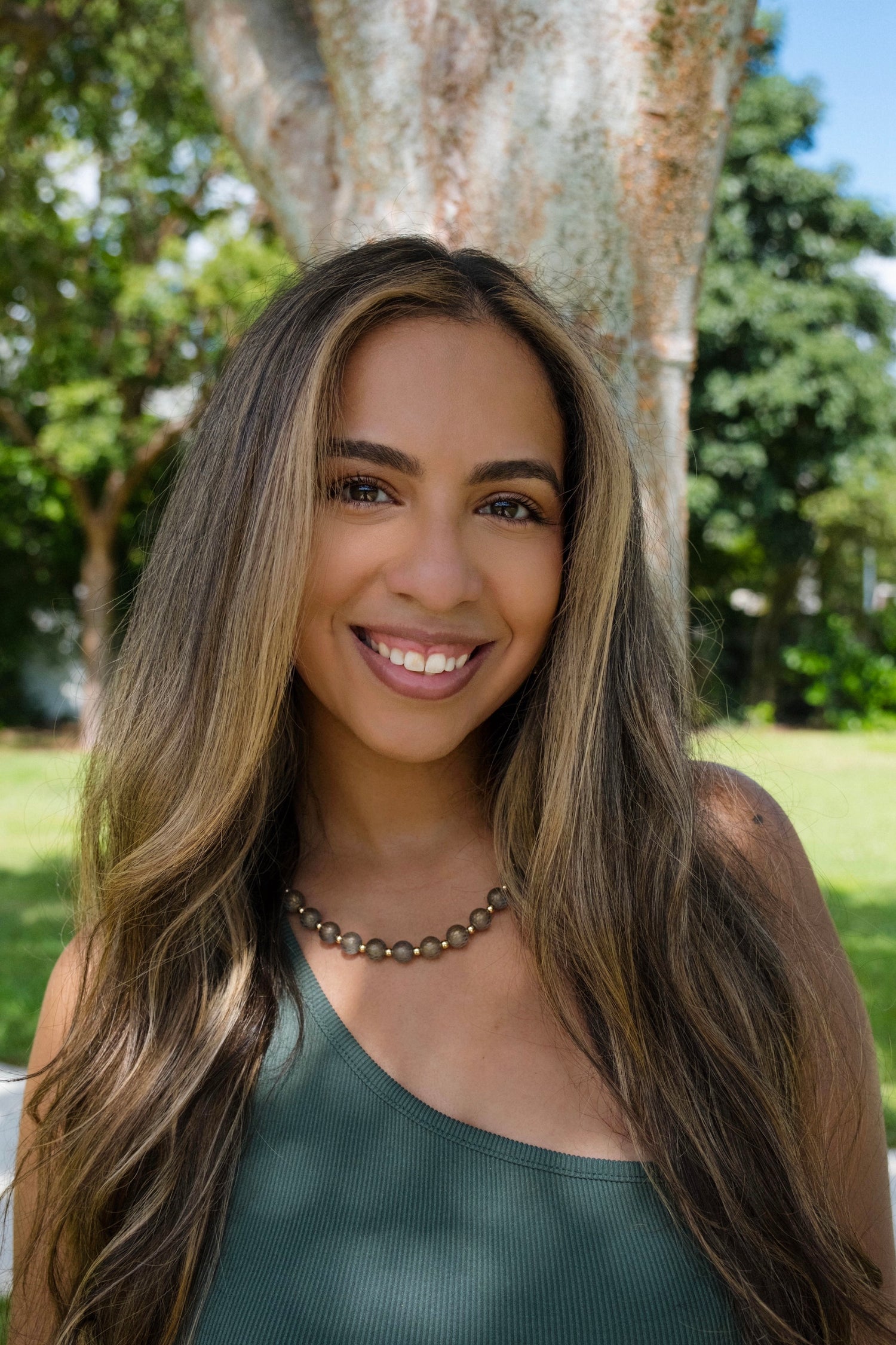 Woman with long brown hair smiling outdoors with trees in the background