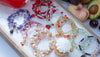 Colorful beaded bracelets on a white surface with fruits in the background
