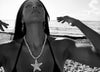 A black and white lifestyle photograph of a woman on a beach, wearing a pearl necklace with a starfish pendant and dangling earrings, looking upwards toward the sun with her arms outstretched.