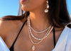 A woman wearing layered pearl and gold necklaces with a teardrop pendant, matching drop earrings, and a black bikini top against a bright blue sky background.