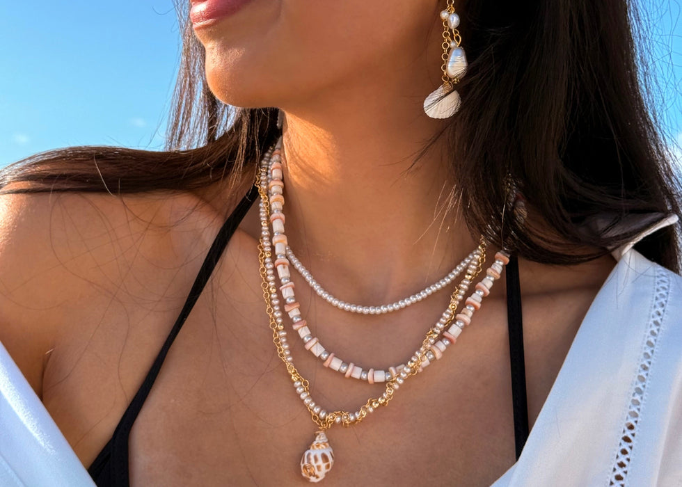 A woman wearing layered pearl and gold necklaces with a teardrop pendant, matching drop earrings, and a black bikini top against a bright blue sky background.