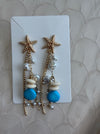 A pair of gold-toned dangle earrings with starfish charms, white shell beads, and bright blue beads, displayed on a white card over a textured beige fabric background.