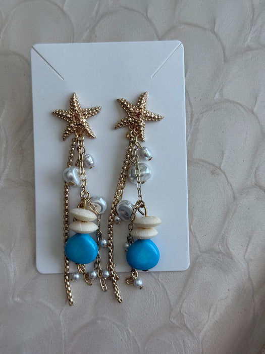 A pair of gold-toned dangle earrings with starfish charms, white shell beads, and bright blue beads, displayed on a white card over a textured beige fabric background.