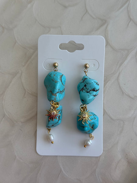 Turquoise earrings with gold accents and pearl-like beads on a white card against a beige textured background.