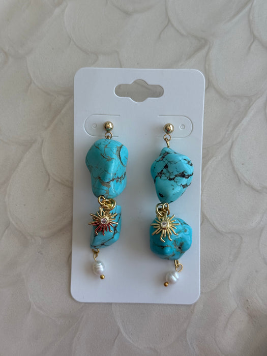 Turquoise earrings with gold accents and pearl-like beads on a white card against a beige textured background.